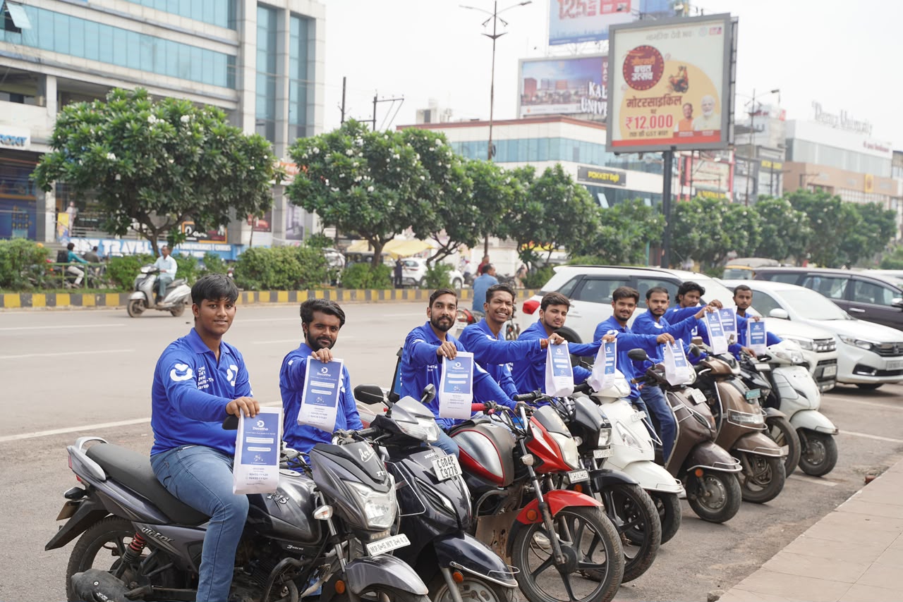 Docatho riders lined up on bikes, holding branded packets toward the camera